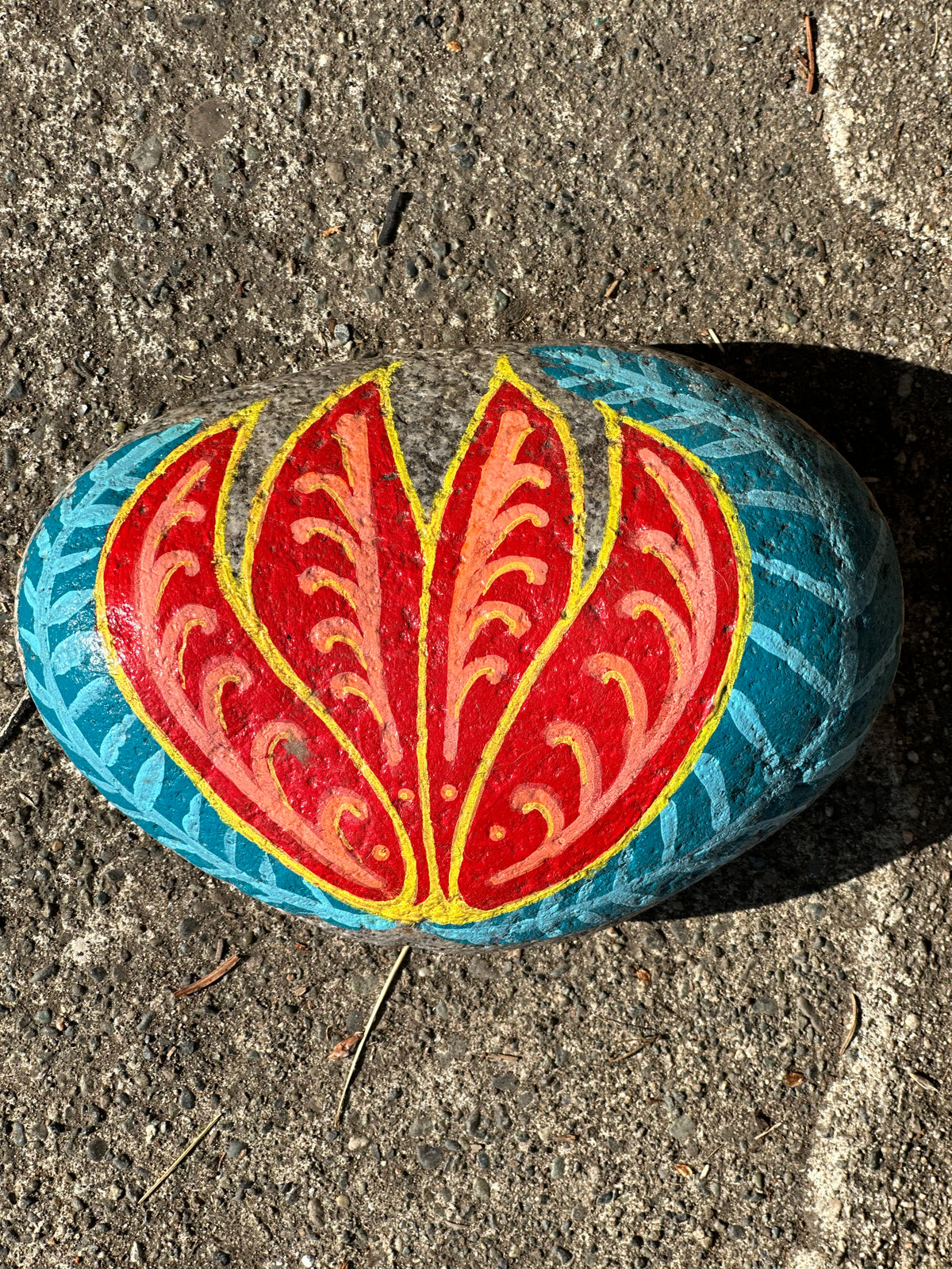 A hand-painted decorative rock featuring a red lotus flower design with yellow and orange details, set against a blue patterned background, photographed on a gray concrete surface.