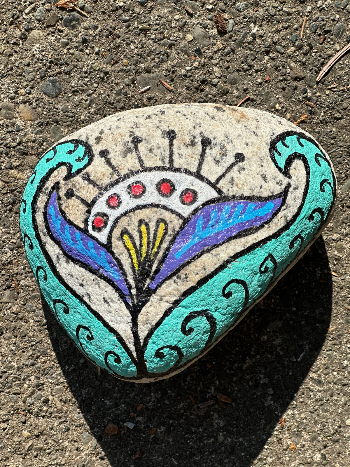 A hand-painted decorative river stone with a stylized floral and crown motif in turquoise, purple, blue, black, red, and yellow, photographed outdoors on a textured concrete surface.