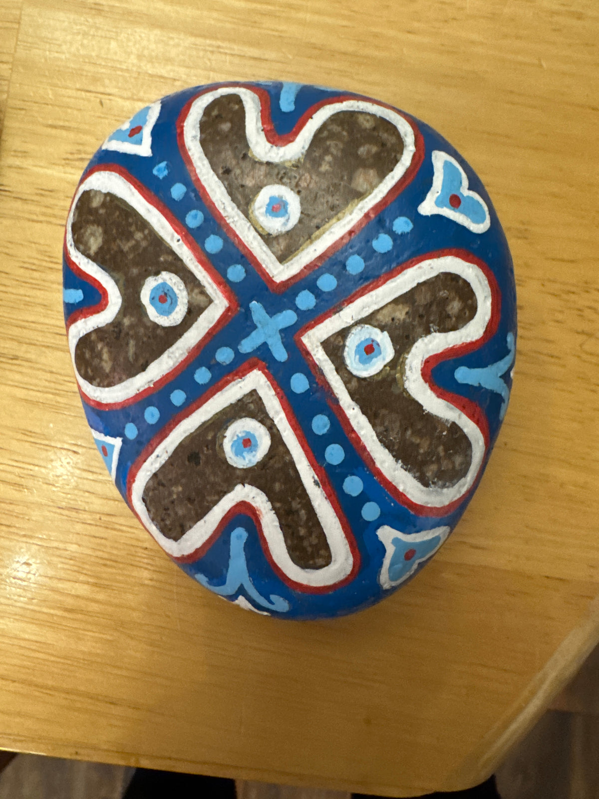 A hand-painted decorative stone featuring a symmetrical cross design in blue, white, red, and brown, resting on a light wood tabletop.