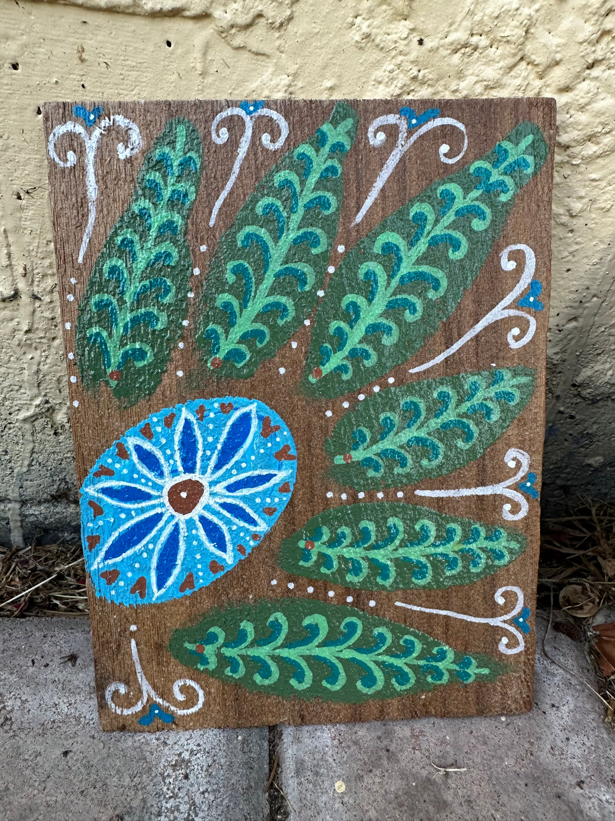 A rectangular wooden plaque featuring a hand-painted folk art design with green and teal leaves, a blue flower motif, and white scroll accents on a brown wood background, leaning against a textured beige wall.