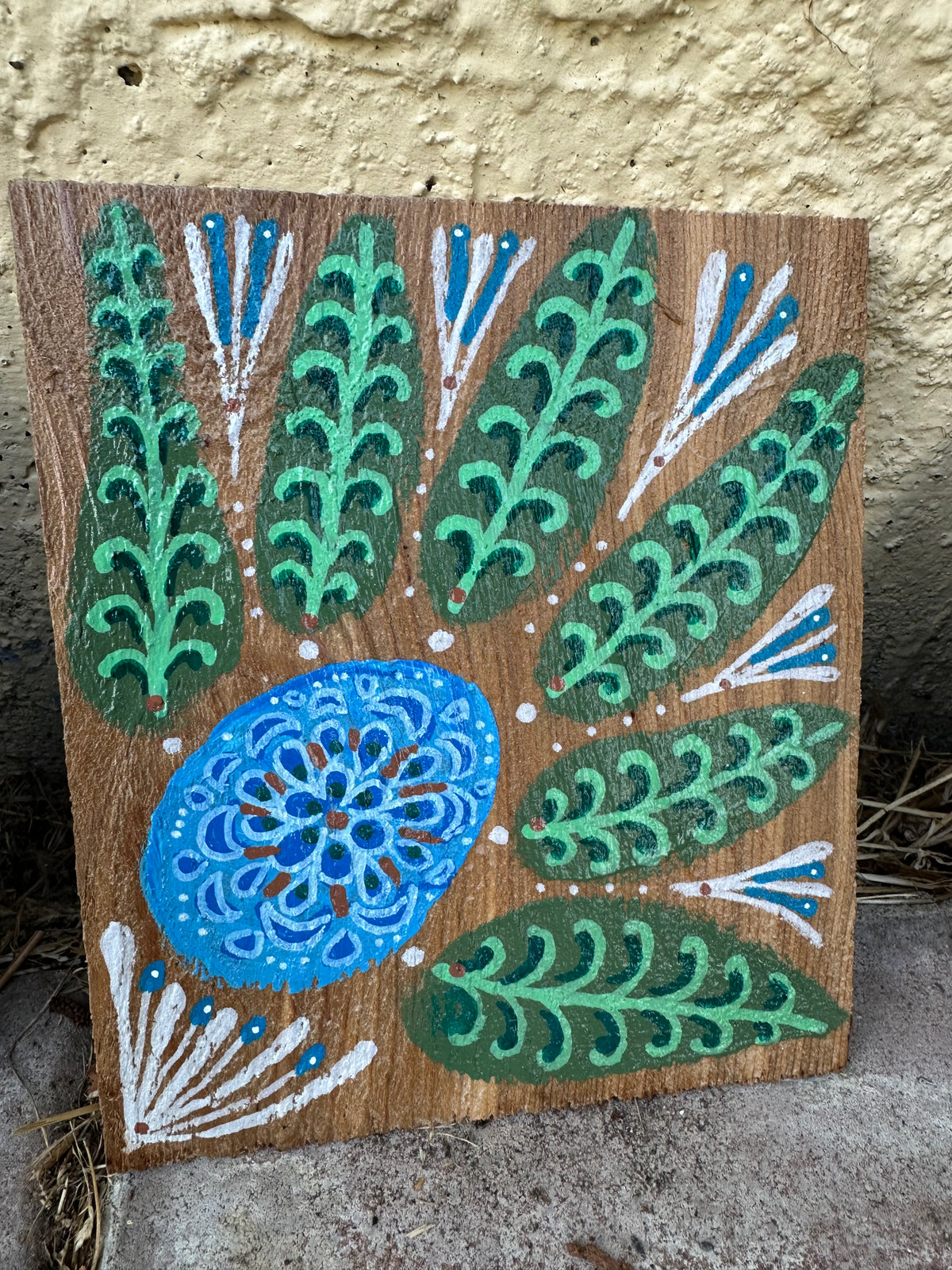 A square wooden art panel featuring a hand-painted folk art design with green leafy motifs, a blue mandala-style oval, and white decorative accents on a natural wood background, leaning against a textured stucco wall.