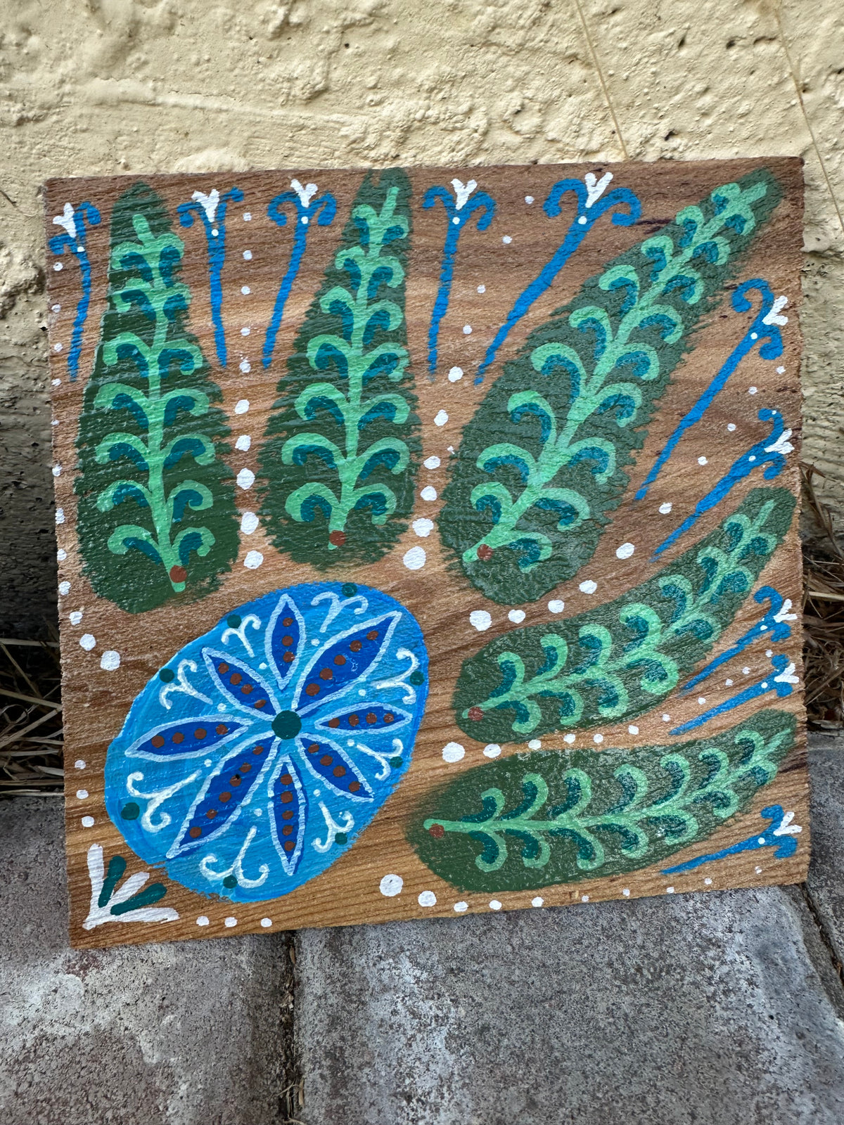 A square wooden decorative plaque featuring a hand-painted folk art design with green and blue stylized leaves and a blue floral motif on a brown wood background, leaning against a textured beige wall and gray stone surface.