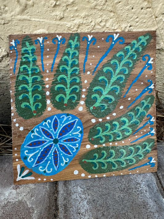 A square wooden decorative plaque featuring a hand-painted folk art design with green and blue stylized leaves and a blue floral motif on a brown wood background, leaning against a textured beige wall and gray stone surface.