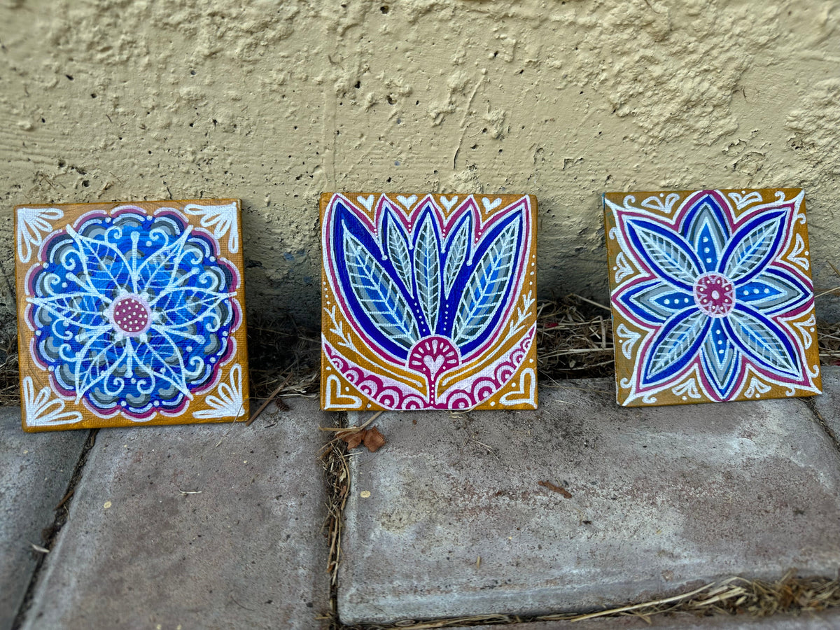 Three square hand-painted ceramic tiles with intricate floral and geometric patterns in blue, purple, pink, and white, displayed on a stone surface against a textured beige stucco wall.