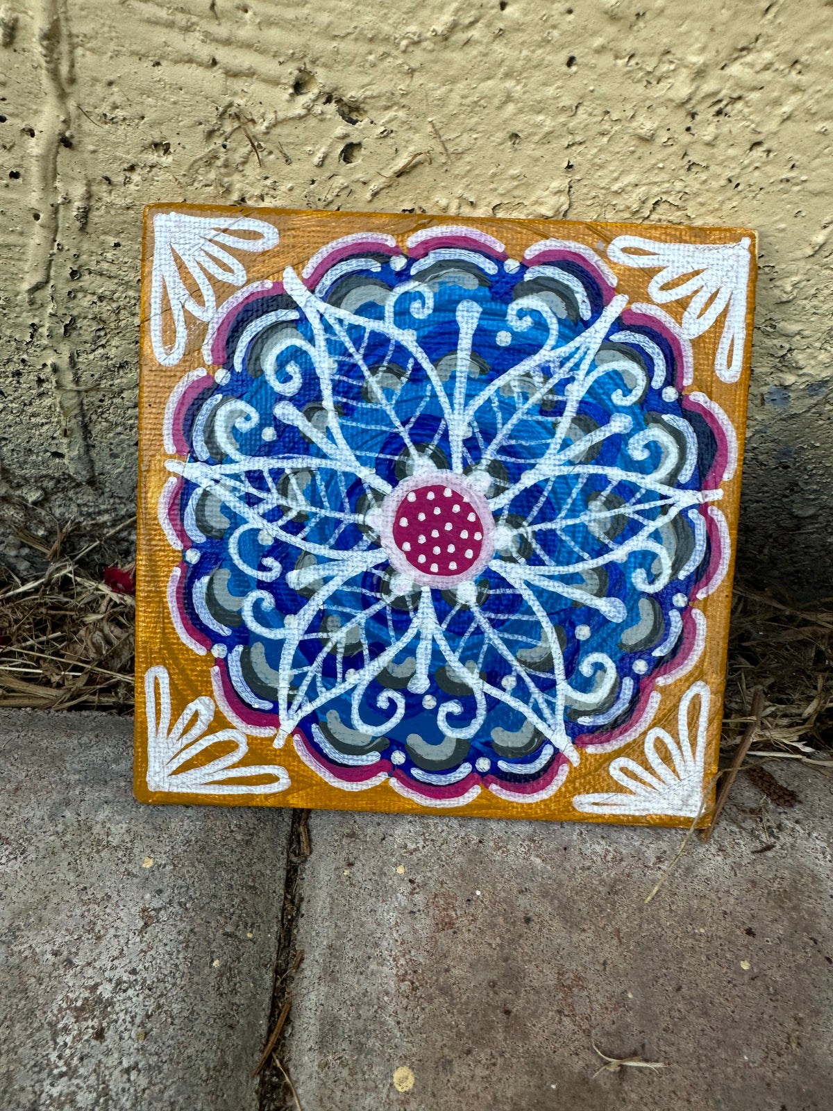 A square canvas painting featuring a colorful mandala design, displayed outdoors against a textured beige stucco wall and concrete ground.