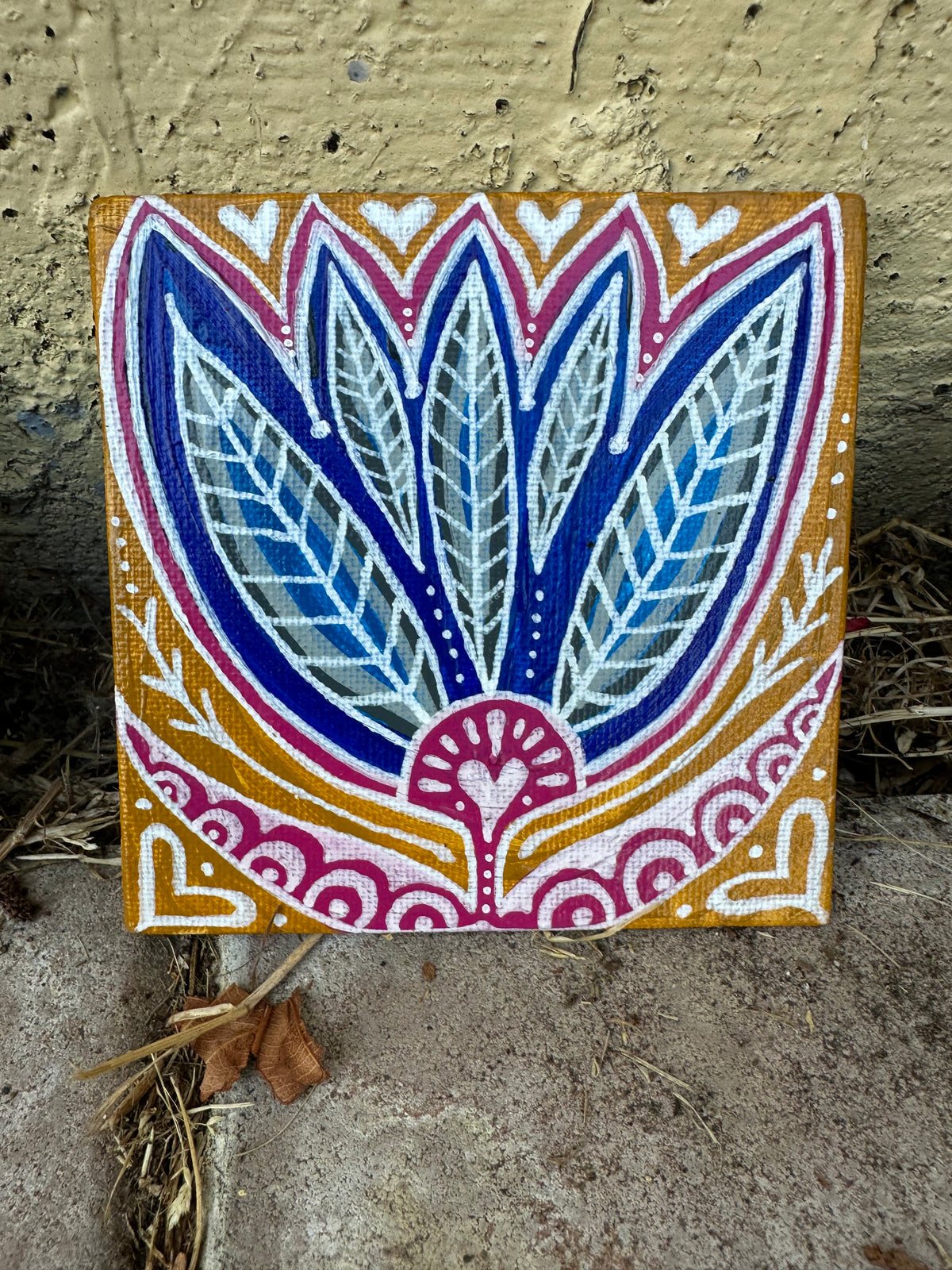 A square canvas painting featuring a symmetrical lotus flower design in blue, pink, white, and gold, photographed leaning against a textured concrete wall on the ground.
