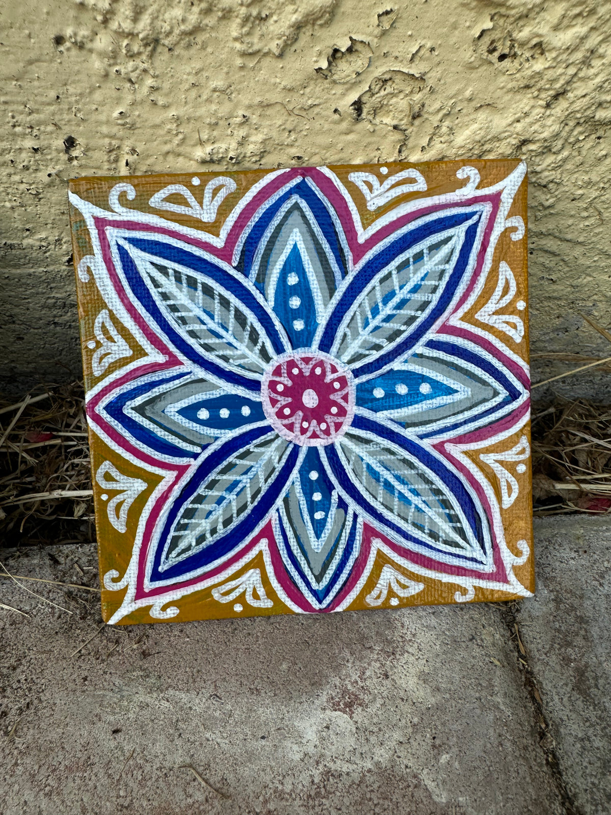 A square decorative tile or canvas featuring a colorful mandala-style floral design, photographed leaning against a textured beige wall and concrete ground.