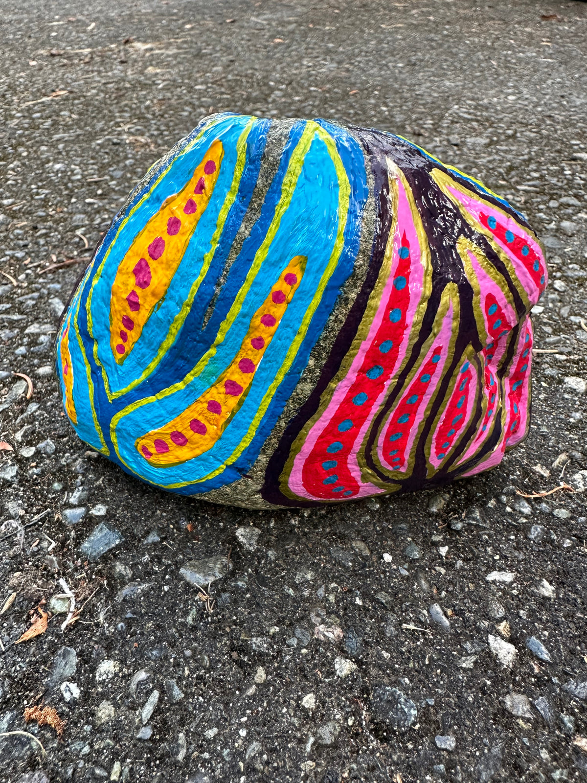 A round decorative rock painted with bold abstract swirls and stripes in bright blue, pink, yellow, black, and red, photographed outdoors on dark asphalt pavement.