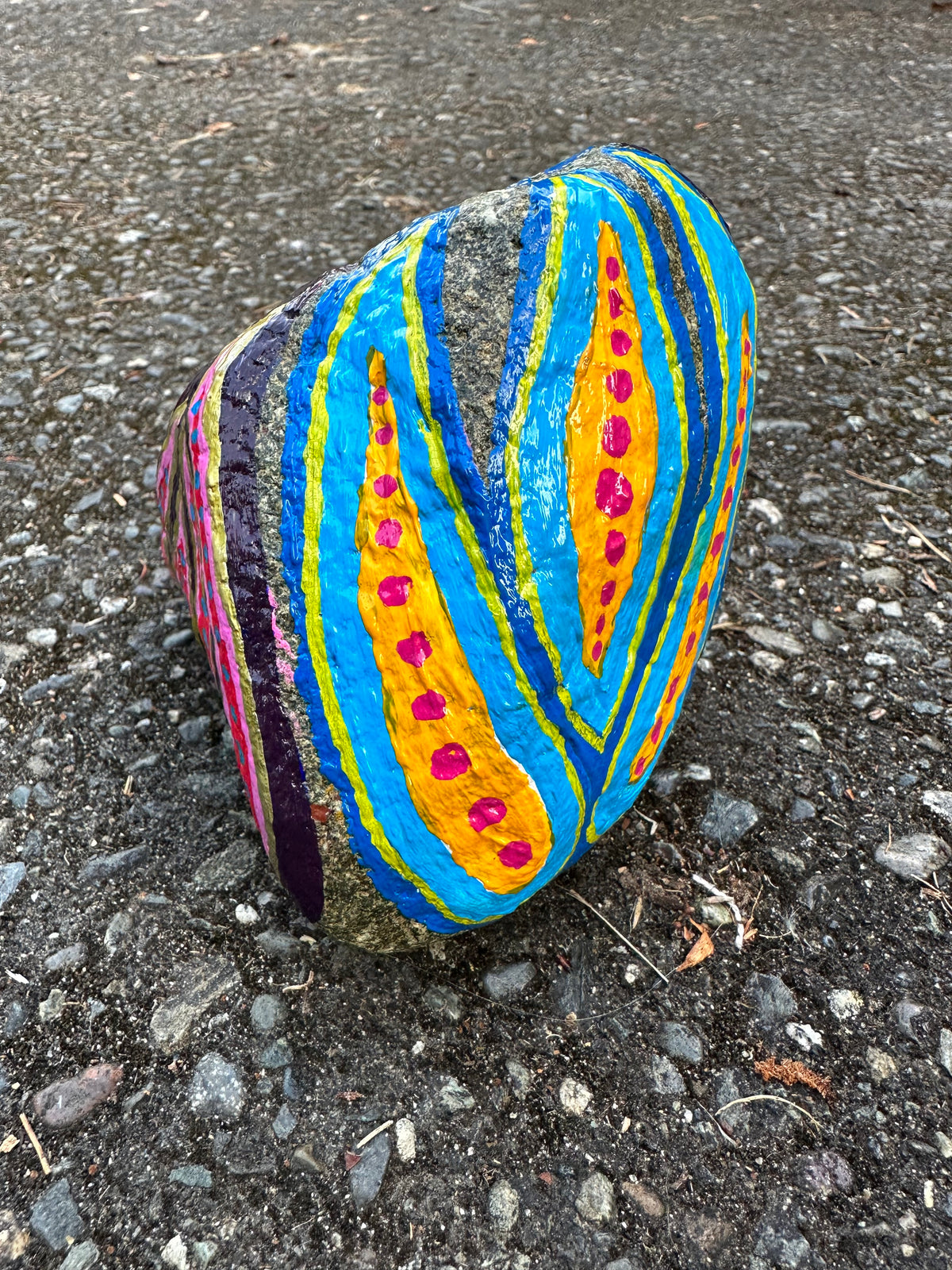 A hand-painted decorative rock sculpture featuring bold vertical stripes in blue, yellow, orange, pink, and purple, standing on a gravel surface.