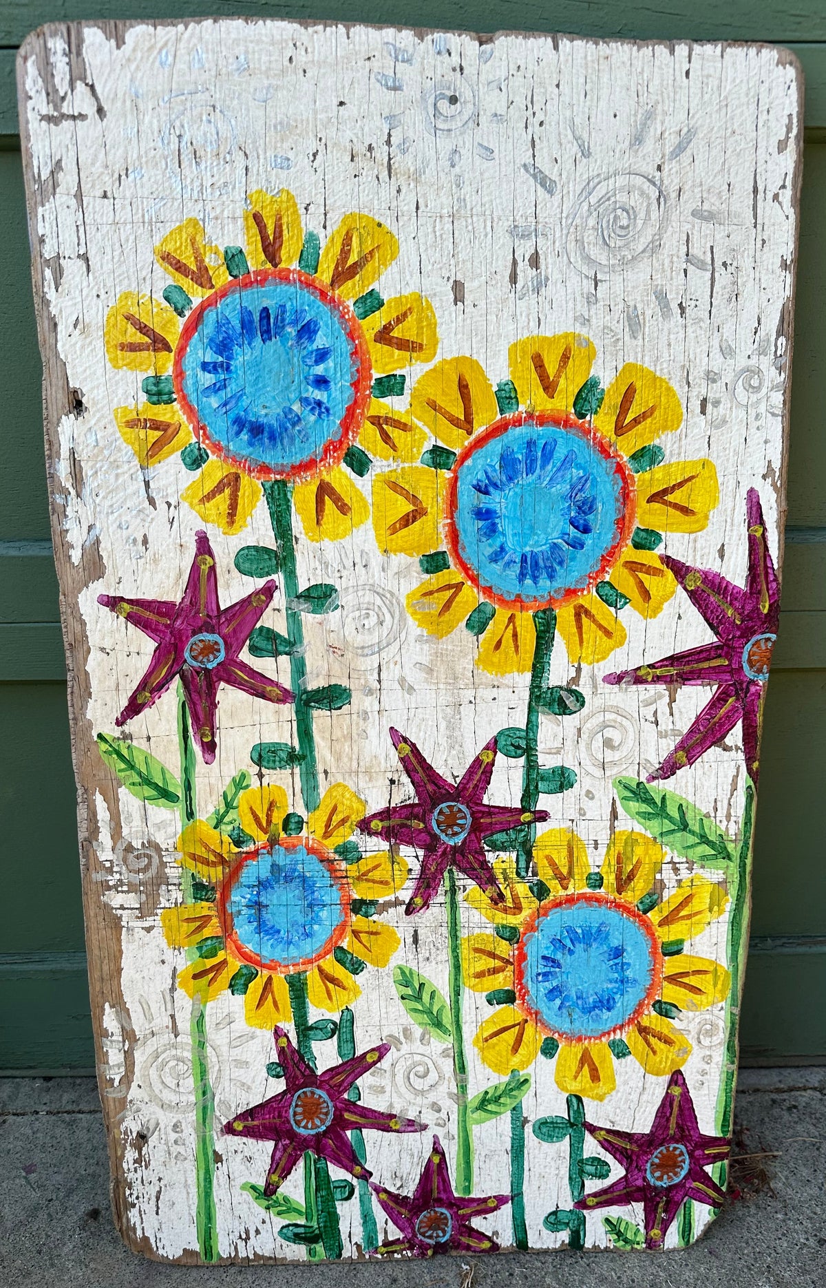 A rustic rectangular wooden sign with a hand-painted floral design featuring yellow sunflowers with blue centers, green stems, and purple star-shaped flowers on a distressed white background, leaning against a green wooden wall.
