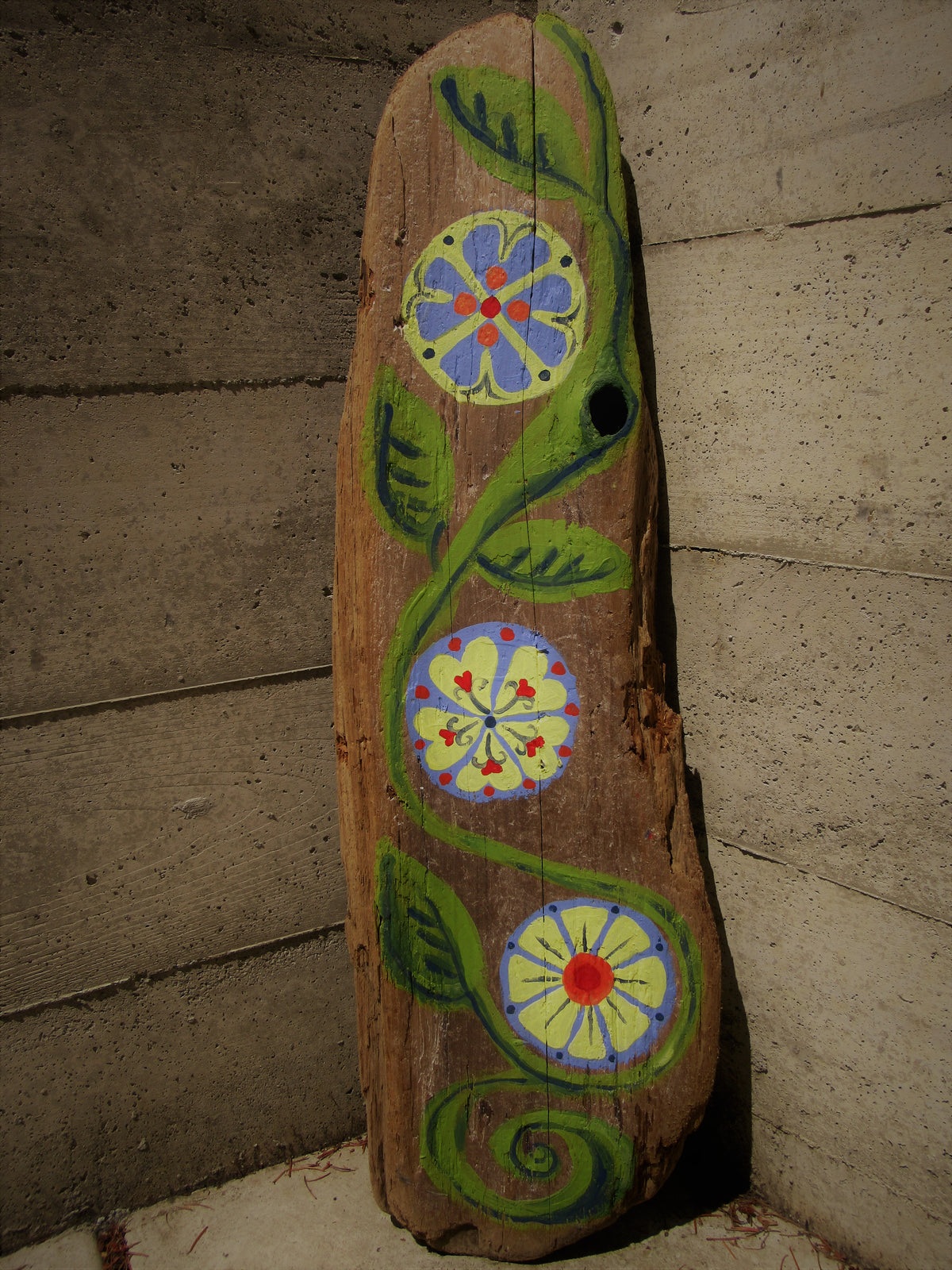 Folk art flowers painted on driftwood by Lone Peep: a green vine with three round blue, yellow and red blooms.