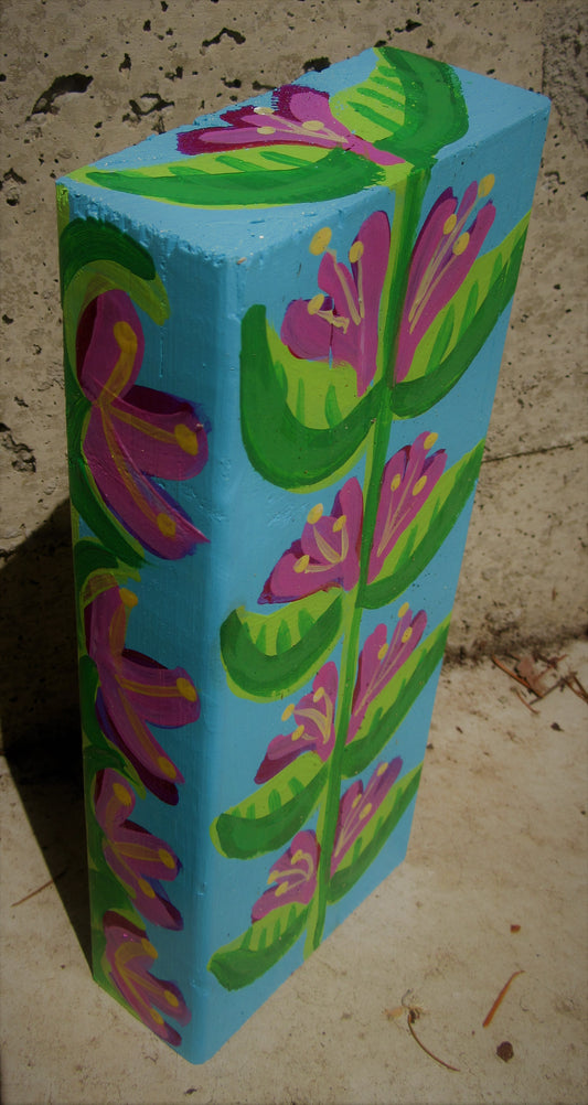 Folk art flowers painted on a wooden block by Lone Peep: magenta blooms and green leaves on bright blue.