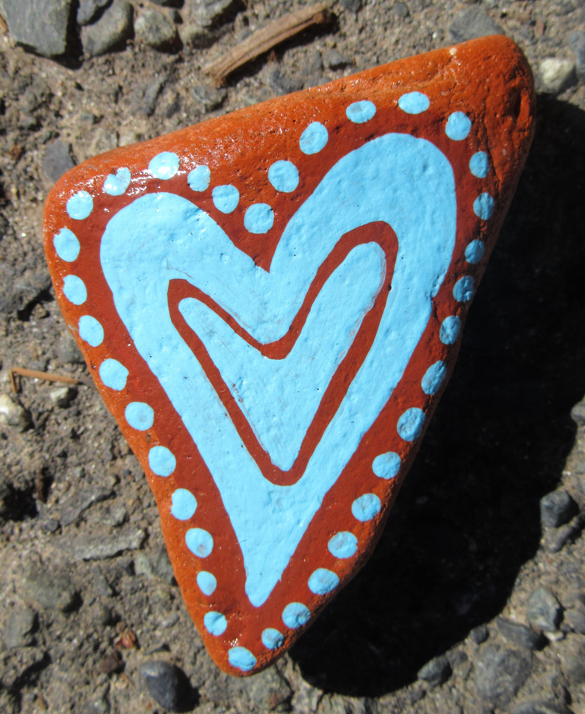 Painted stone by Lone Peep: terracotta stone with a light blue heart and dotted border.