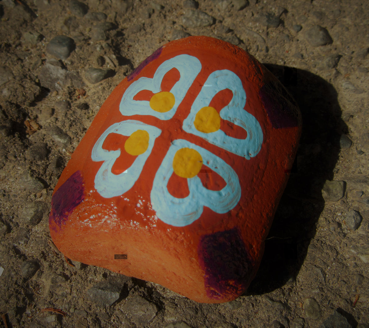 Painted stone by Lone Peep: terracotta stone with a light blue four-petal flower and yellow dots.