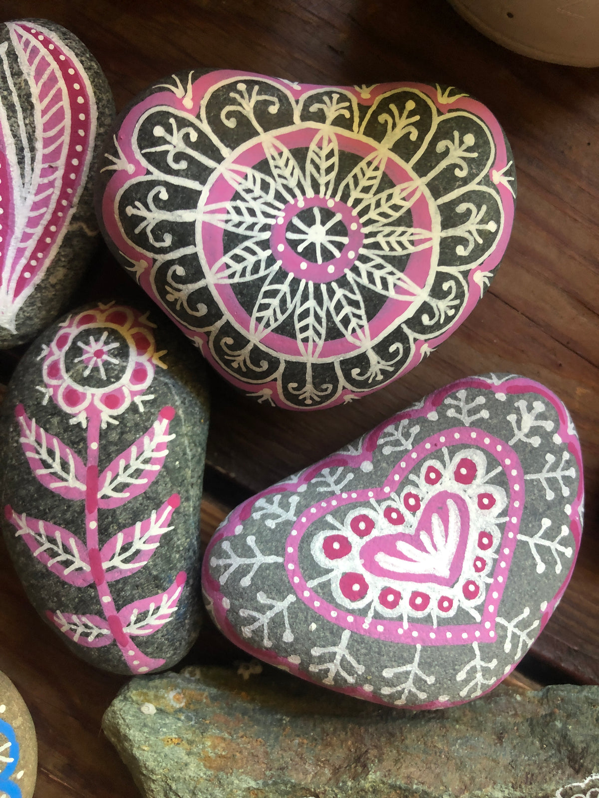 Three hand-painted decorative stones with intricate mandala and floral patterns in pink, white, and black, arranged on a dark wooden surface.