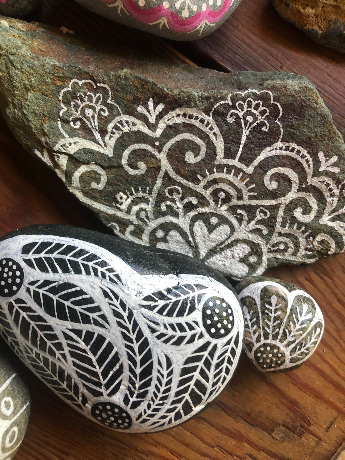 Decorated garden stones with intricate hand-painted white and black patterns, including a heart-shaped stone, a large flat rock, and a small scalloped stone, arranged on a wooden surface.