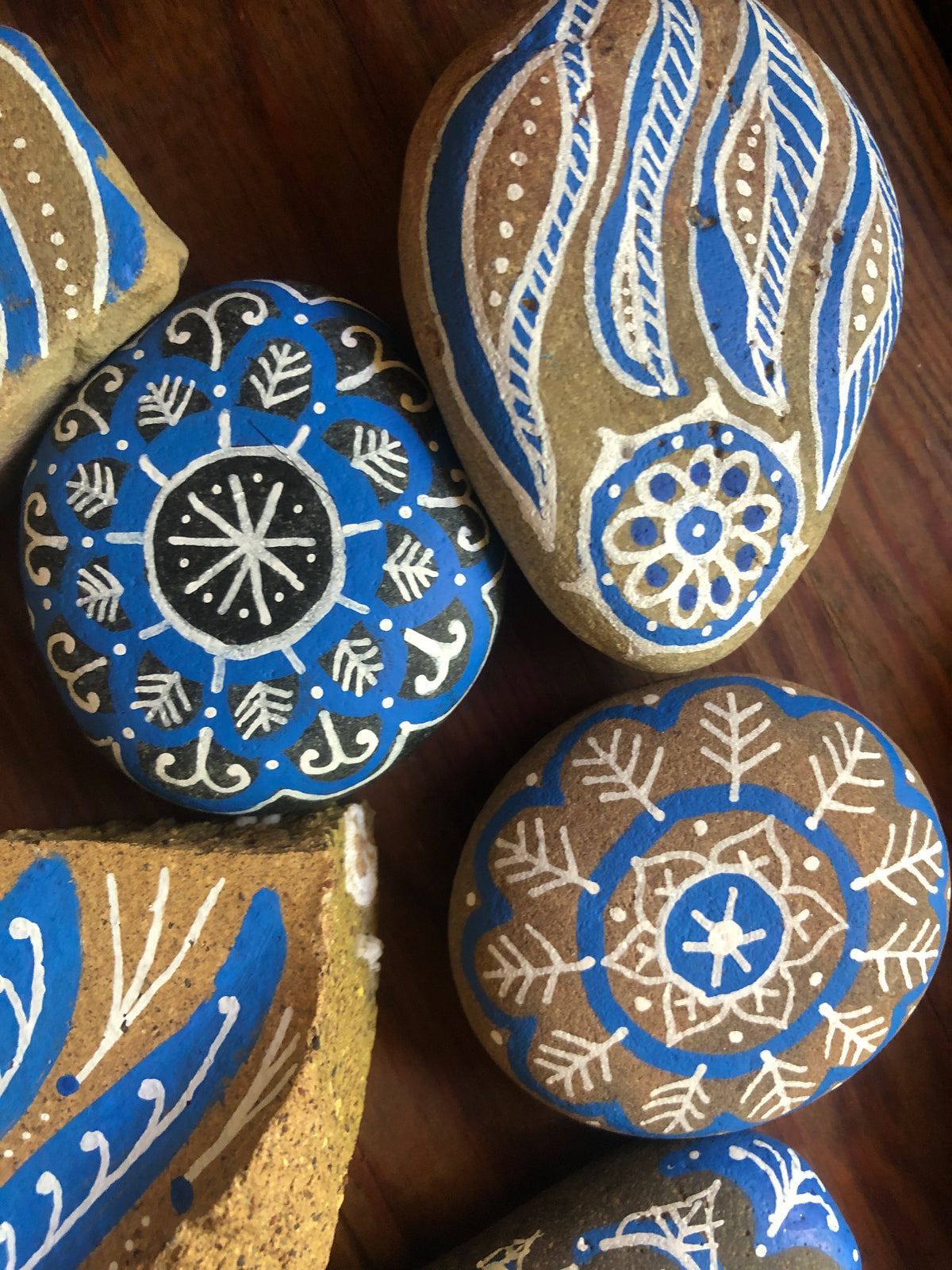 A close-up of several hand-painted decorative rocks with intricate blue, white, and tan patterns arranged on a dark wooden surface.